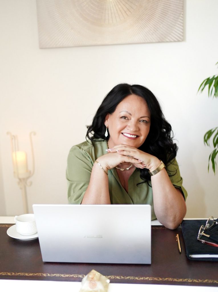 Life and relationship coach Bernadett smiling warmly at her desk with a laptop, ready to help clients on their journey to self-discovery and soul harmony.